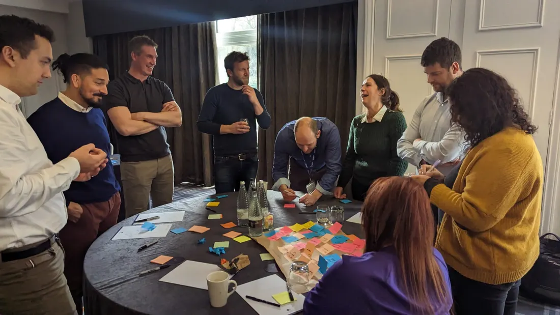A team standing around a round table covered in sticky notes during a workshop, several people laughing